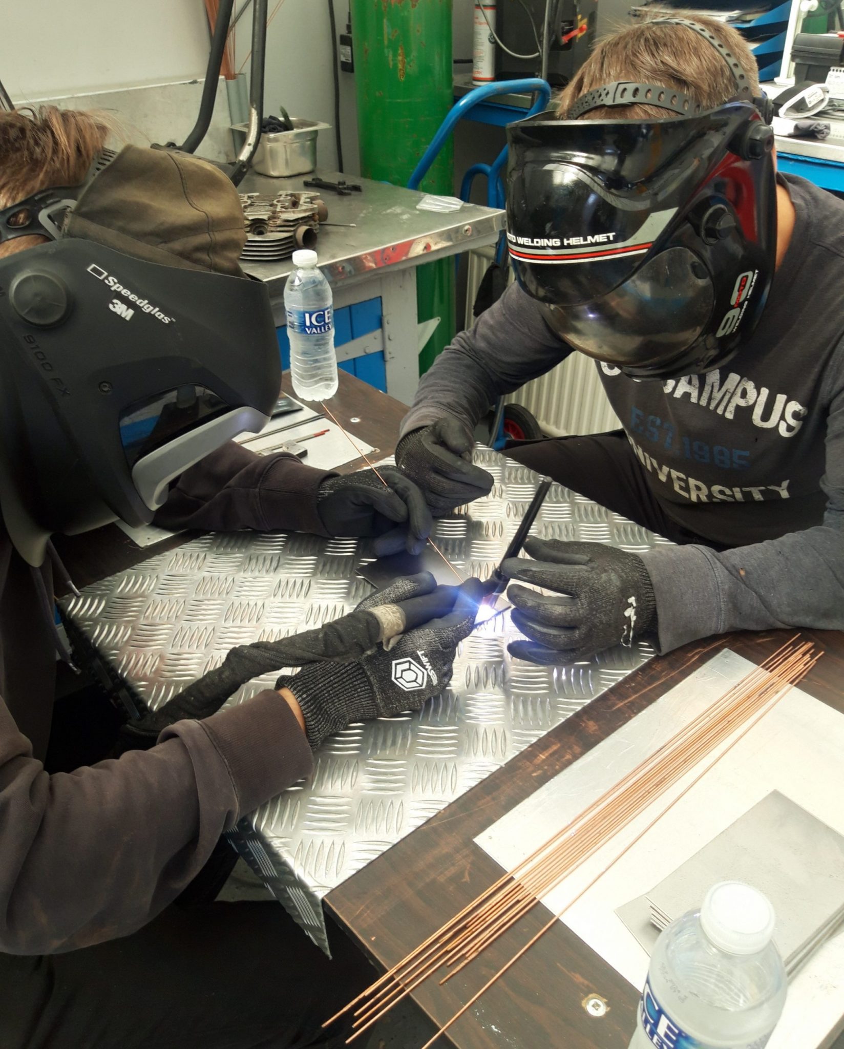 Two people wearing welding helmets and gloves are welding a metal sheet on a table, with welding tools and water bottles nearby.