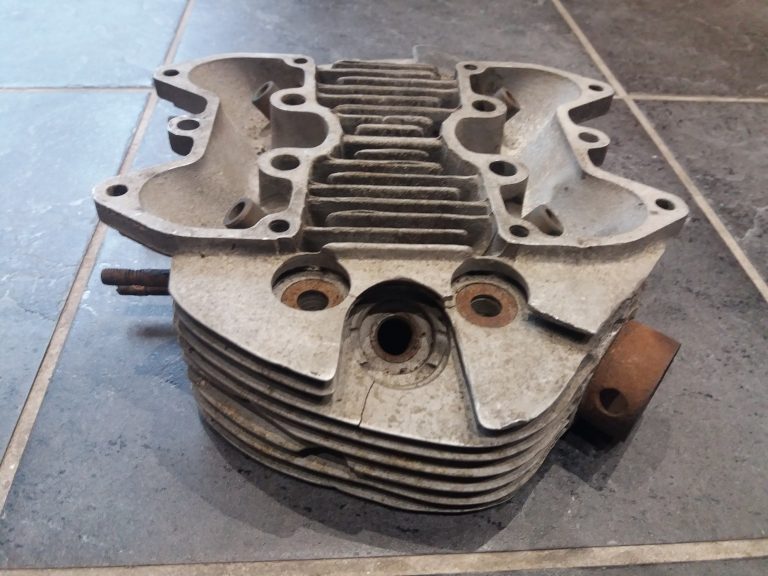 A used metal engine cylinder head with visible cooling fins rests on a tiled floor.