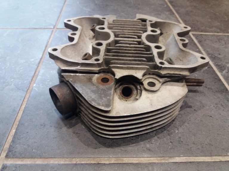 A metal engine cylinder head with cooling fins is placed on a tiled floor, showing various ports and bolt holes.