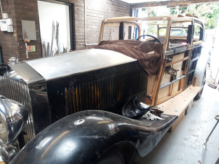 A vintage car undergoing restoration in a garage, with exposed wooden framework and metal body panels partially installed.