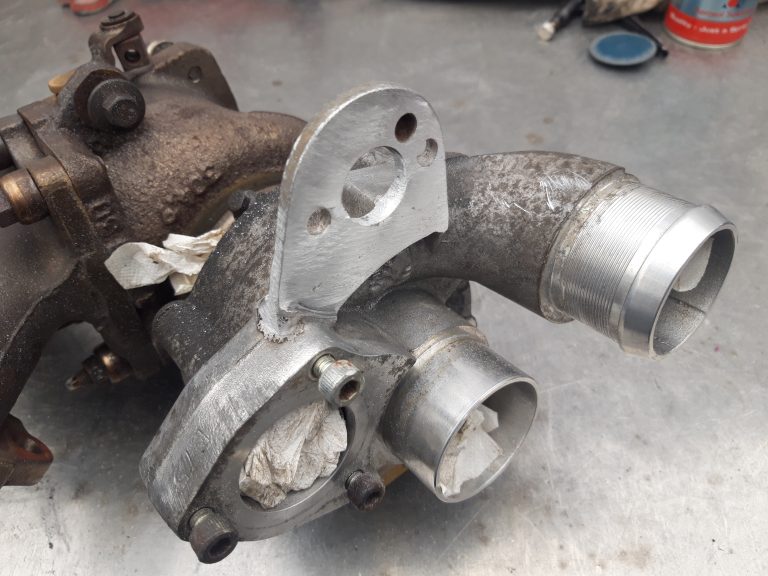 A close-up view of a metal automotive turbocharger component with machined surfaces and paper plugs in the openings, resting on a metal workbench.