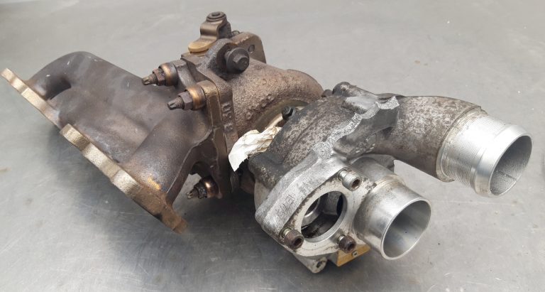 A used automotive turbocharger with an exhaust manifold, placed on a metallic surface.