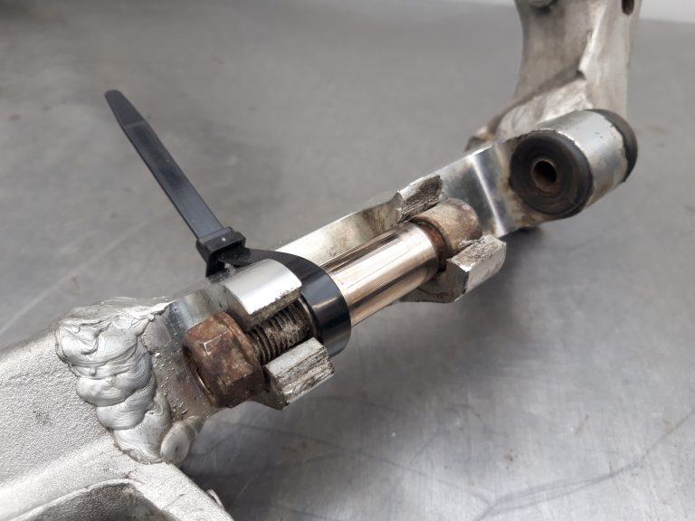 Close-up of a metal mechanical part with a bolt, cylindrical pin, and a black zip tie securing components together on a gray surface.