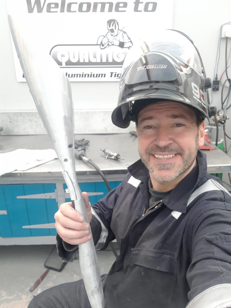 A man in work overalls and a welding helmet smiles while holding a metallic propeller-shaped object in a workshop. A sign reading "Welcome to Qualtig Aluminium Tig" is behind him.
