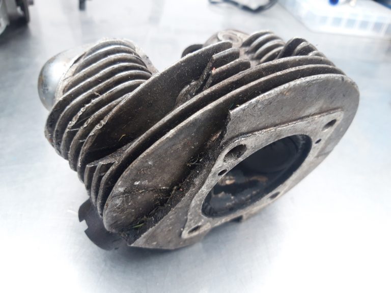 A close-up of a used, dirty aluminium cylinder head with cooling fins, placed on a metallic surface—ideal for showcasing aluminium cylinder head repairs.