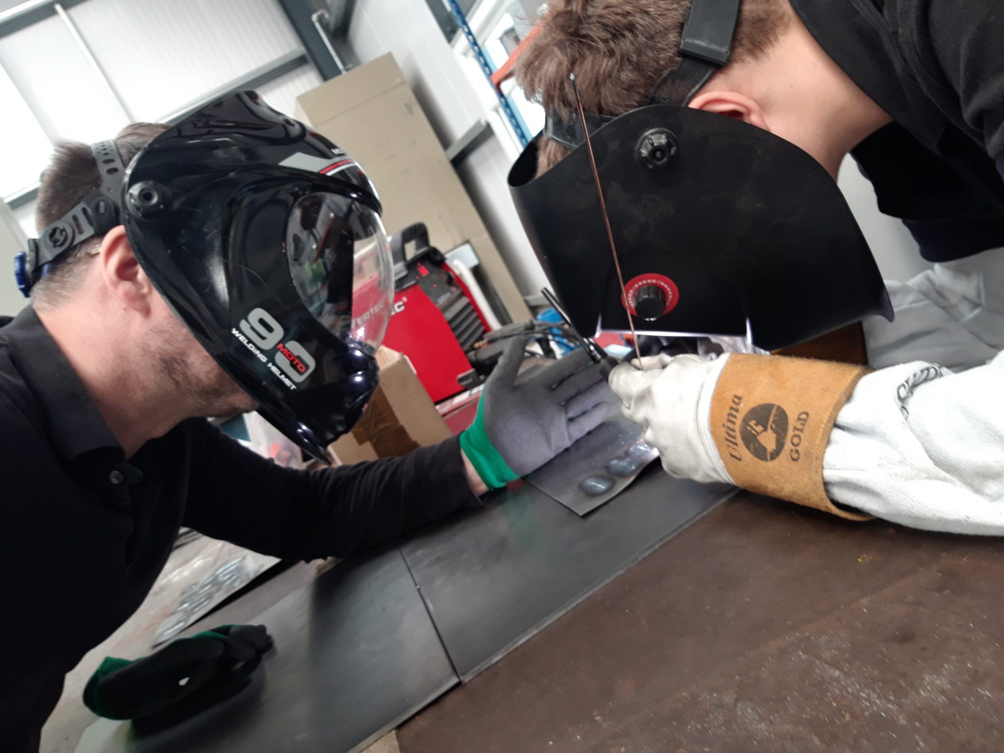 Two people wearing welding helmets work together on a metal welding project at a workshop table.