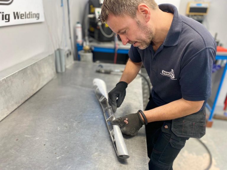 A man wearing gloves works on a metal pipe at a stainless steel workbench in a workshop.