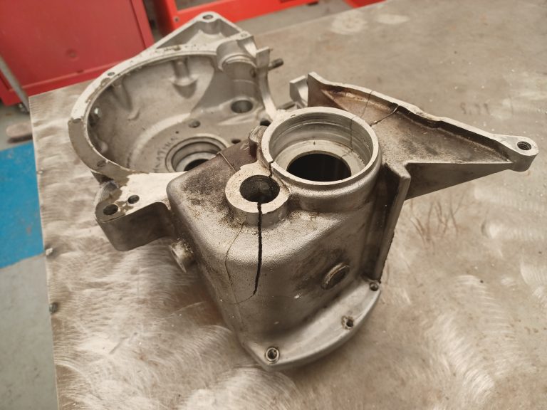 A cracked metal engine casing sits on a scratched workbench in a workshop setting, awaiting specialist aluminium repair work.