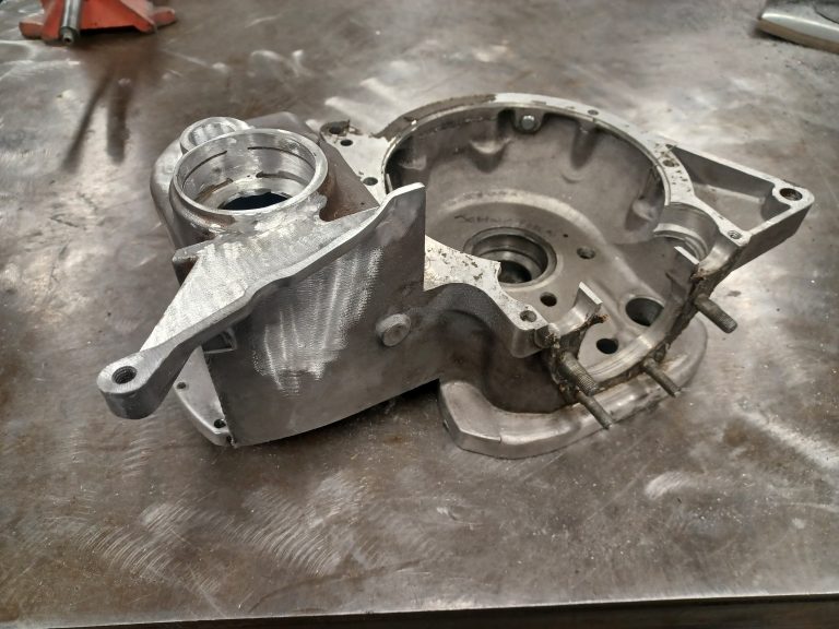 A metal engine housing component with visible machining marks, indicative of specialist aluminium repair work, rests on a scratched metal workbench.