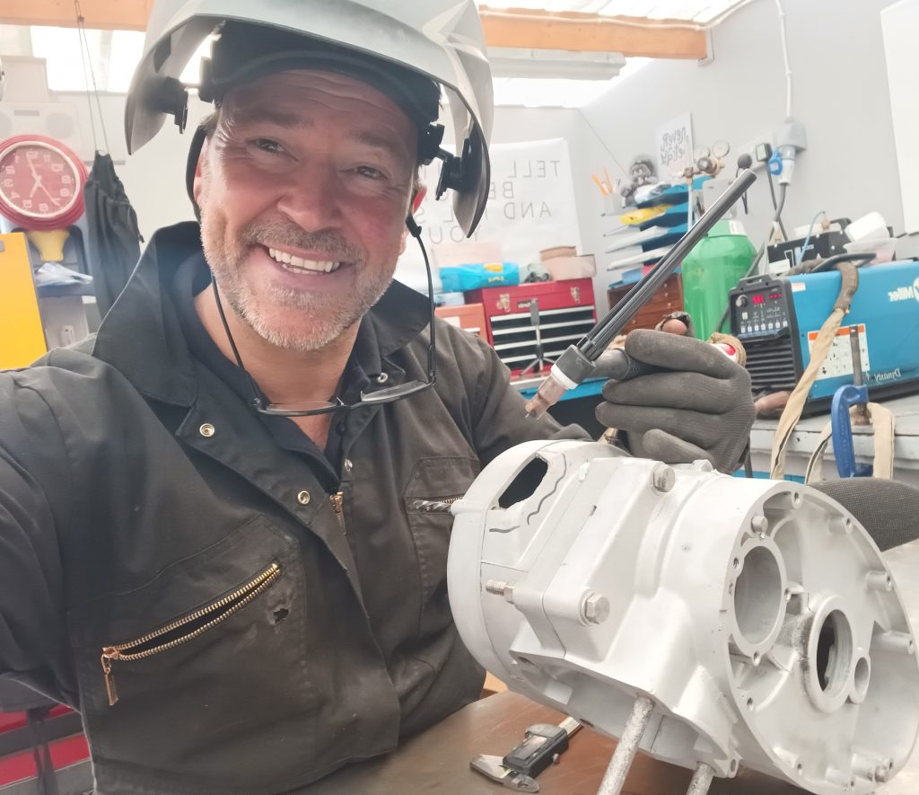 Man wearing protective gear smiles at the camera while holding a tool and working on a metal machine part during Hands-On Tig & Mig Welding & Restoration in Coventry.
