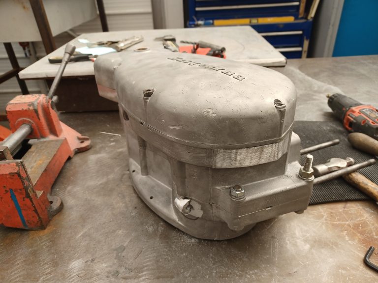 A metal gearbox housing sits on a workbench surrounded by tools including a drill, pliers, and a vise.