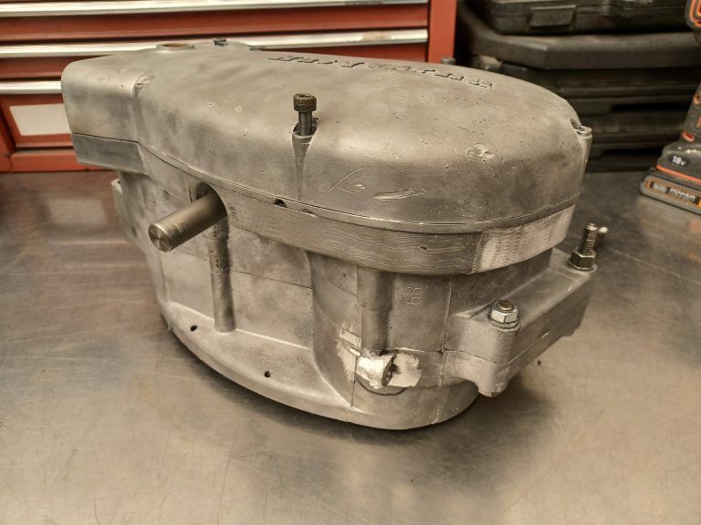An aluminum motorcycle engine casing with bolts and a protruding shaft rests on a metal workbench in a workshop setting.