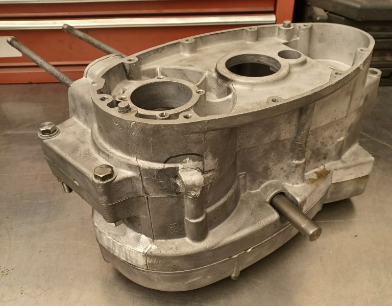Aluminum motorcycle engine crankcase with visible mounting bolts and shafts, placed on a metal workbench with a red toolbox in the background.