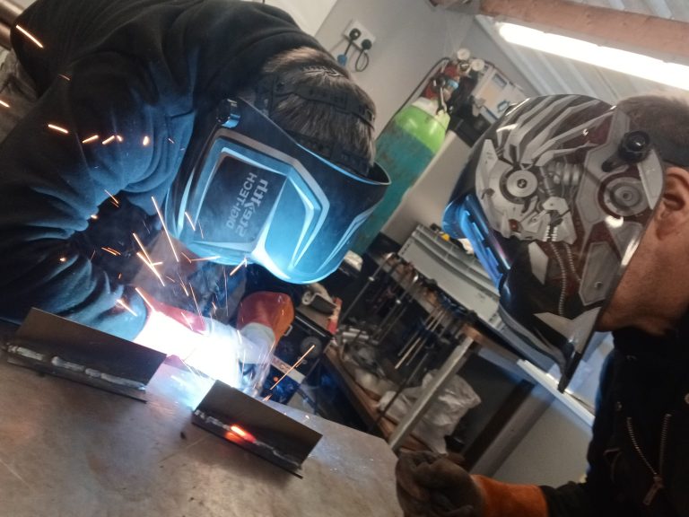 Two welders in protective gear working on metal components, sparks flying, demonstrating TIG welding techniques in a workshop setting for classic vehicle repairs.