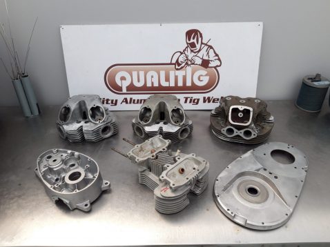 Seven metal engine parts are arranged on a table in front of a “Qualitig” sign, with welding rods and a spool of wire visible in the background.
