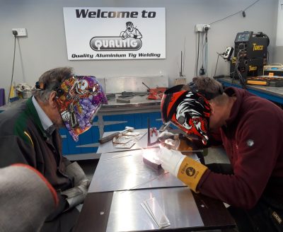Two people in protective welding masks work on a metal piece in a workshop, with a sign reading “Welcome to Quality Aluminium Tig Welding” in the background. Two people in protective welding masks work on a metal piece in a workshop, with a sign reading "Welcome to Quality Aluminium Tig Welding" in the background.